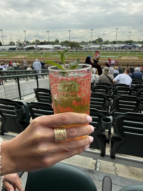 Mint julep held up at trackside, hand wearing the 18K yellow gold diamond fashion ring with the racetrack in the background